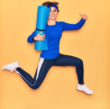 Genç ve güzel sporcu kadın mutlu gülümsüyor. Yüzünde tebessümle yoga minderini tutarak zıplıyorum. Tamam işareti yapıyorum. Başparmaklarımla izole edilmiş sarı arka plan üzerinde..