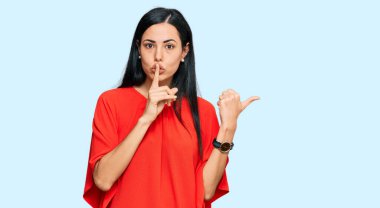 Gündelik giysiler giyen genç ve güzel bir kadın sessiz olmak istiyor. Parmakları dudaklarında, elleri yana doğru işaret ediyor. sessizlik ve gizli konsept. 