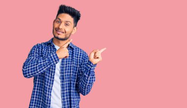 Yakışıklı Latin Amerikalı rahat gömlekli genç adam gülümsüyor ve kameraya bakıyor iki eliyle ve parmaklarıyla kenarı işaret ediyor.. 