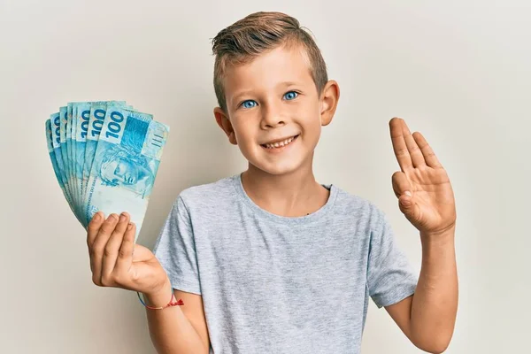Uroczy Biały Dzieciak Trzymający 100 Brazylijskich Banknotów Robi Znak Palcami — Zdjęcie stockowe