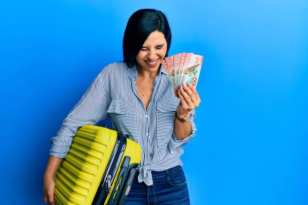 Young caucasian woman holding cabin bag and new zealand dollars banknotes smiling and laughing hard out loud because funny crazy joke.
