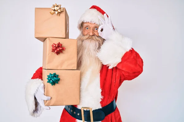 Old senior man with grey hair and long beard wearing santa claus costume holding presents smiling happy doing ok sign with hand on eye looking through fingers