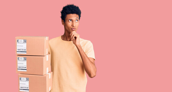 Young african american man holding delivery package serious face thinking about question with hand on chin, thoughtful about confusing idea 