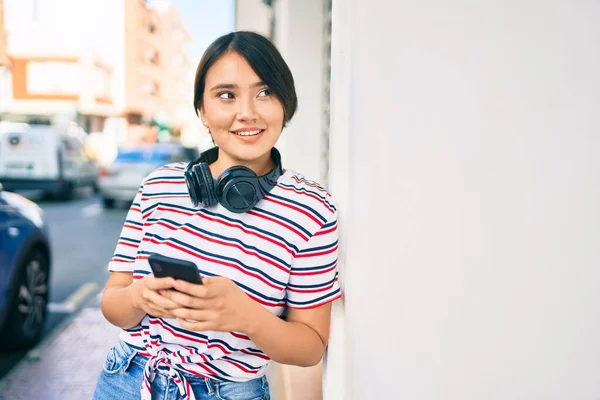 Young latin girl smiling happy using smartphone and headphones at the city