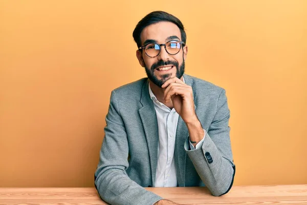 Young hispanic man with beard wearing business clothes sitting on the table looking confident at the camera smiling with crossed arms and hand raised on chin. thinking positive.