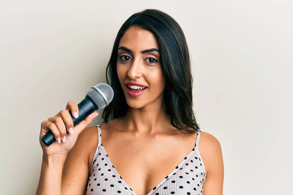 Beautiful hispanic woman singing song using microphone looking positive and happy standing and smiling with a confident smile showing teeth