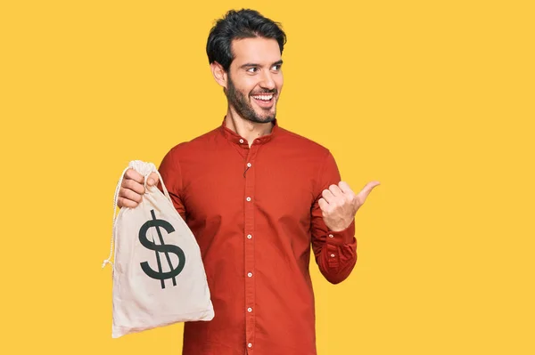 Young Hispanic Man Holding Dollars Bag Pointing Thumb Side Smiling — Stock Photo, Image