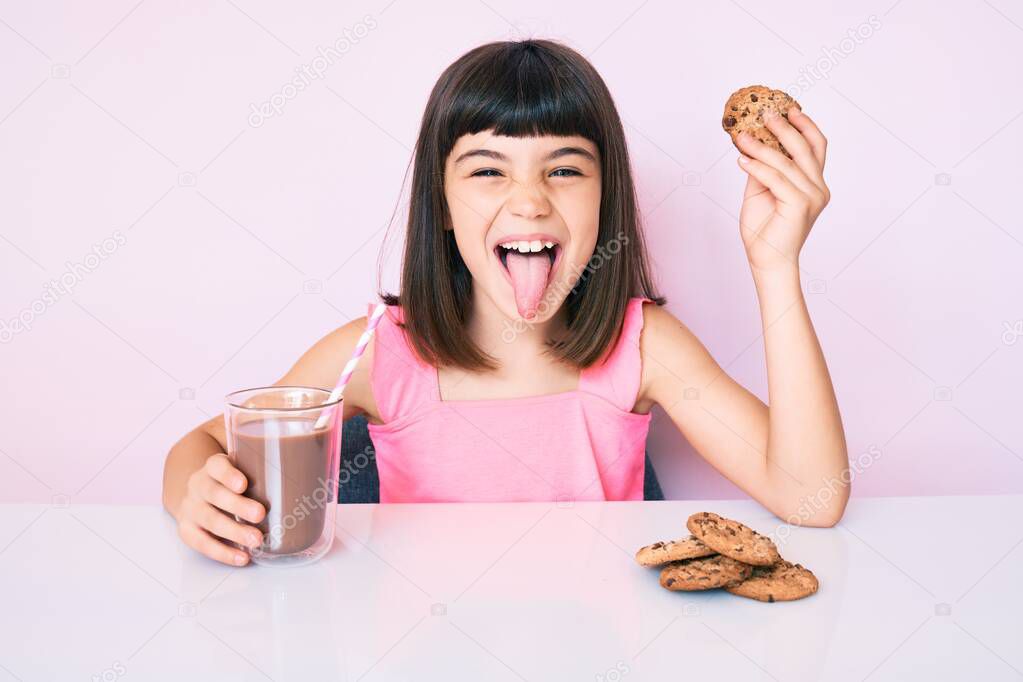 Young little girl with bang sitting on the table having breakfast sticking tongue out happy with funny expression. 