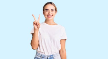 Genç, güzel, sarışın, günlük beyaz tişörtler giyen, iki numaralı parmaklarıyla işaret eden, kendine güvenen ve mutlu bir kadın.. 