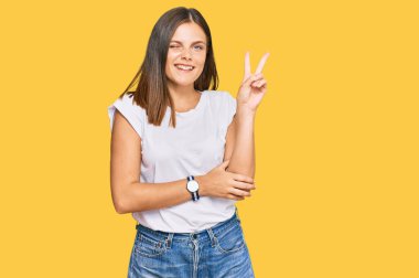 Gündelik beyaz tişört giyen, kameraya gülümseyen ve zafer işareti yapan beyaz bir kadın. İki numara.. 