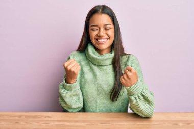 Güzel İspanyol kadın, gündelik giysiler içinde masada mutlu ve heyecanlı bir şekilde oturuyor. Kolları havada, gülümseyerek ve başarı için çığlık atarak kazanan jestler yapıyor. kutlama konsepti. 
