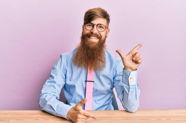 Genç İrlandalı kızıl adam iş gömleği ve kravat takıyor neşeli bir şekilde masada oturuyor yüzünde bir gülümsemeyle mutlu ve doğal bir ifadeyle elini ve parmağını yana kaldırıyor. 