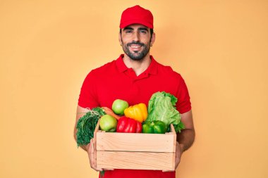 Yakışıklı, sakallı, üniformalı, elinde teslimat yemeği tutan mutlu ve havalı bir gülümsemeyle İspanyol bir adam. diş gösterme. 
