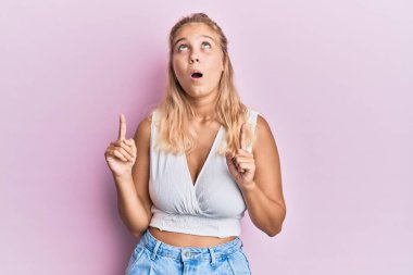 Gündelik kıyafetler giyen genç sarışın kız şaşırmış ve şaşırmış bir şekilde yukarı bakıyor ve parmaklarıyla işaret ediyor ve kollarını kaldırıyor.. 