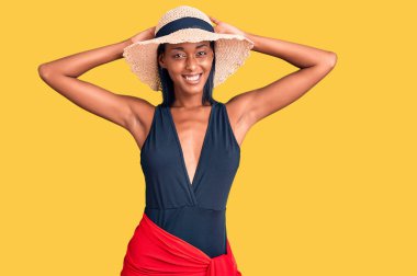 Genç Afro-Amerikalı kadın mayo giyiyor ve yaz şapkası takıyor. Rahatlıyor ve esniyor. Kolları ve elleri başının arkasında. 