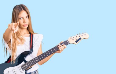 Güzel, beyaz, sarı saçlı, elektro gitar çalan, kamerayı işaret eden, kendine güvenen, ciddi görünen bir kadın. 