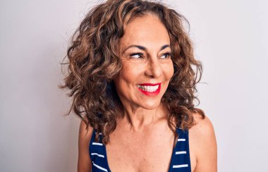 Orta yaşlı, esmer, çizgili tişört giyen güzel bir kadın beyaz arka planda durup yan tarafa bakıyor, rahat bir görünüm takınıyor ve kendine güvenen bir gülümseme sergiliyor..