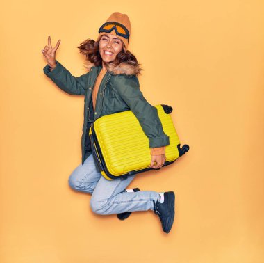 Kışlık giysiler giyen genç, güzel Latin kadın mutlu gülümsüyor. Yüzünde gülümsemeyle kamara çantası tutarak zıplıyor izole edilmiş sarı arka planda zafer işareti yapıyor.