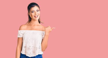 Genç, güzel bir kadın, günlük kıyafetler giyiyor, gülümsüyor, gülümsüyor, gülümsüyor ve başparmağıyla yan tarafa bakıyor.. 