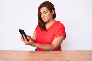 Orta yaşlı esmer kadın akıllı telefon kullanıyor. Masada oturuyor. Düşünme tavrı takınıyor. 