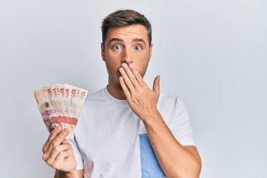 Elinde 10 Kolombiya pesosu tutan yakışıklı beyaz adam ağzını eliyle kapatıyor, şaşırmış ve hata yapmaktan korkuyor. şaşırmış ifade 