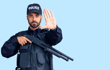Polis üniforması giyen genç İspanyol adam elinde pompalı tüfekle dur işareti yapıyor ciddi ve kendinden emin bir ifadeyle, savunma hareketi yapıyor. 