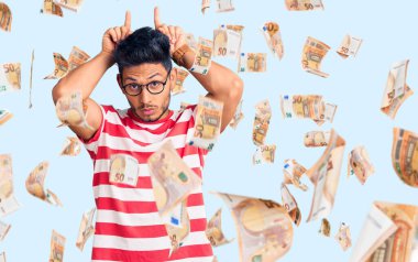 Yakışıklı Latin Amerikalı genç adam, günlük kıyafetler ve gözlük takıyor parmağıyla boynuz gibi komik hareketler yapıyor.