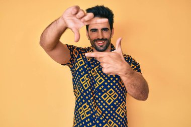 80 'lerin disko tarzı tişörtü giyen sakallı yakışıklı İspanyol adam gülümsüyor ve elleri ve parmakları mutlu yüzlü olan bir çerçeve yapıyor. yaratıcılık ve fotoğrafçılık kavramı. 