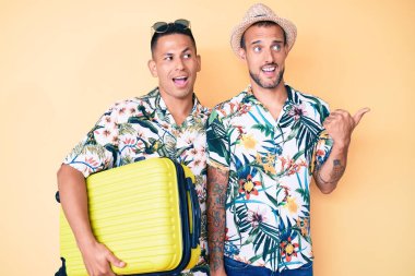 Genç eşcinsel iki adam ellerinde bavullarıyla yaz tatiline çıkıyorlar. Başparmağını kaldırıp gülümsüyorlar. Ağızları açık. 