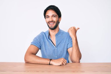 Yakışıklı İspanyol bir adam, günlük giysiler giymiş, masaya oturmuş gülümsüyor, gülümsüyor ve başparmağıyla yan tarafa bakıyor.. 