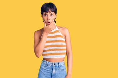 Kısa saçlı, günlük kıyafetler giyen genç esmer kadın elleri çenesinde, inanamama, şaşkınlık ve şaşkınlık dolu bir ifade takınıyor. 