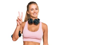 Spor kıyafetleri giyen ve kulaklık takan genç sarışın kadın iki numarayı gösterip işaret ederken kendinden emin ve mutlu bir şekilde gülümsüyor.. 