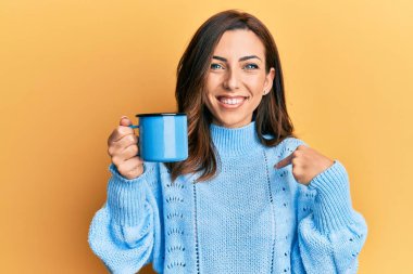 Genç esmer kadın elinde bir fincan kahveyle kendini gösteren mutlu ve gururlu bir kadını işaret ediyor. 