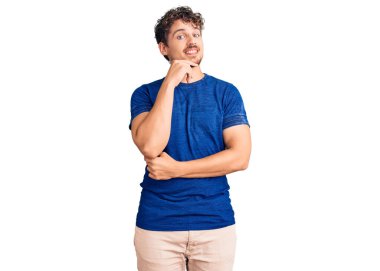 Kıvırcık saçlı, sıradan kıyafetler giyen, kameraya kendinden emin, kollarını kavuşturmuş ve çenesini kaldırmış gülümseyen genç yakışıklı bir adam. Olumlu düşünmek. 