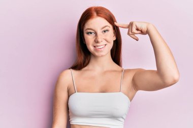 Pembe arka planda duran genç kızıl saçlı kız mutlu bir şekilde elini ve parmağını gösterdi. 