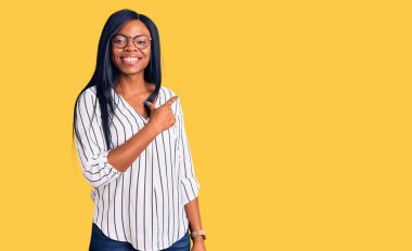 Genç Afrikalı Amerikalı kadın, günlük kıyafetler ve gözlük takıyor. Neşeli, yüzünde bir gülümseme var. Yüzünde mutlu ve doğal bir ifade var. 