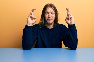 Uzun saçlı yakışıklı beyaz adam masanın üzerinde oturmuş umut ve gözlerin kapalı bir şekilde gülümseyerek işaret ediyor. Şans ve batıl inançlar. 