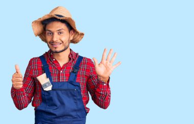 Yakışıklı Latin Amerikalı genç adam, tamirci üniforması giyiyor. Altıncı parmaklarını gösterip gösterirken, kendinden emin ve mutlu bir şekilde gülümsüyor.. 