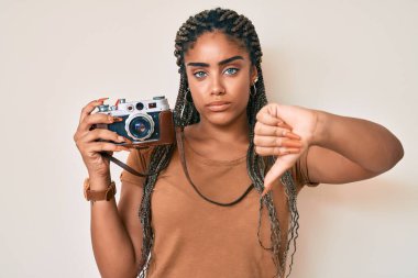 Saçları örgülü, kızgın yüzlü, antika fotoğraf makinesi olan genç bir Afro-Amerikalı kadın. Negatif işaret baş parmakları aşağıda, reddedilme konsepti var. 