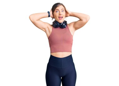 Güzel beyaz kadın spor kıyafetleri giyiyor ve kulaklık takıyor. Çılgınca kulaklık takıyor. Elleri başında, korkmuş ve ağzı açık bir şekilde şoktan korkuyor. 
