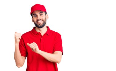 Genç, sakallı, teslimat üniforması giyen, elleri ve başparmakları havada, kendinden emin bir şekilde arkasını gösteren yakışıklı bir adam. 