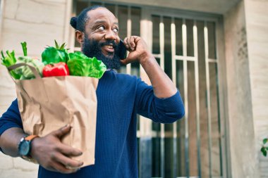 Elinde süpermarketten alınmış kağıt torba sakallı Afrikalı Amerikalı adam telefonda konuşuyor.