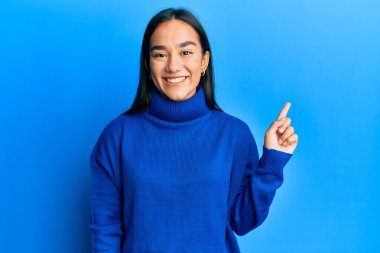 Genç Asyalı kadın, sıradan bir kazak giyiyor. Yüzünde kocaman bir gülümseme var. Parmağıyla kameraya bakıyor.. 