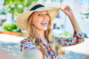 Yaz şapkası takmış, mutlu bir şekilde gülümseyen güzel sarışın bir kadın. Yüzünde gülümsemeyle şehrin caddesindeki kameranın önünde selfie çekerken..