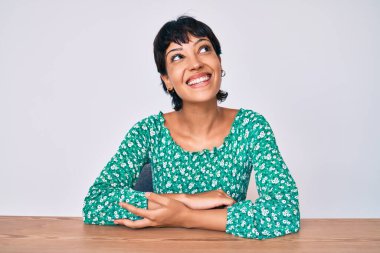 Güzel esmer kadın, gündelik giysiler içinde masada oturuyor, yüzünde gülümsemeyle yan yana bakıyor, doğal bir ifade. Kendinden emin gülen. 