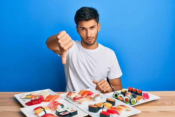 Young handsome man eating sushi sitting on the table looking unhappy and angry showing rejection and negative with thumbs down gesture. bad expression.