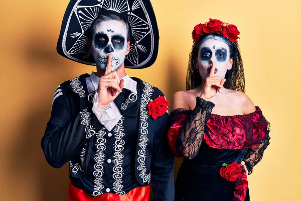 黄色の上に死んだ衣装のメキシコの日を身に着けている若いカップルは 唇に指で静かにするように求めています 沈黙と秘密の概念 — ストック写真