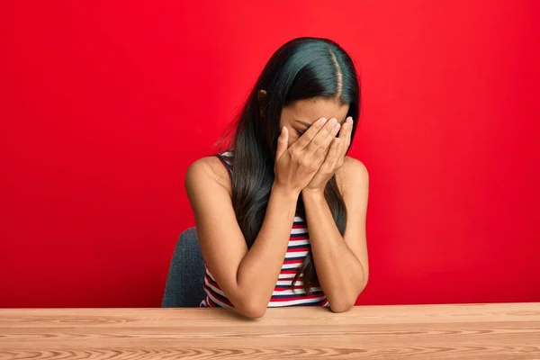 Beautiful hispanic woman wearing casual clothes sitting on the table with sad expression covering face with hands while crying. depression concept.