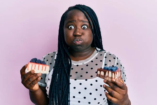 Young black woman with braids holding cheesecakes puffing cheeks with funny face. mouth inflated with air, catching air.