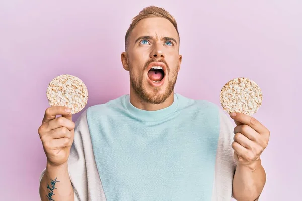 Young Caucasian Man Eating Healthy Rice Crackers Angry Mad Screaming — Φωτογραφία Αρχείου